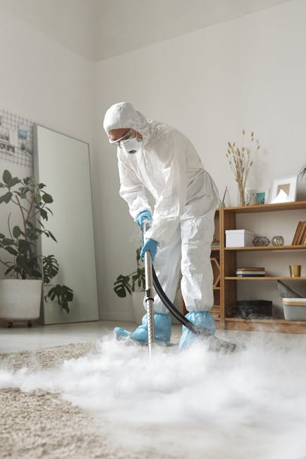 A professional cleaner dressed in full white protective suit, face mask, and gloves performs deep cleaning on a light beige carpet in a modern living room using a steam or dry cleaning machine. The ro