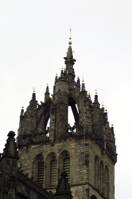 A tall stone church with a pointed spire and a clock, set against a partly cloudy sky with some golden sunlight breaking through. The church is surrounded by lush green trees and a graveyard with neat